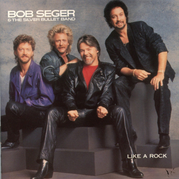 Bob Seger & The Silver Bullet Band album cover for 'Like A Rock', featuring the four band members posed on dark gray steps against a textured blue-gray studio background.
