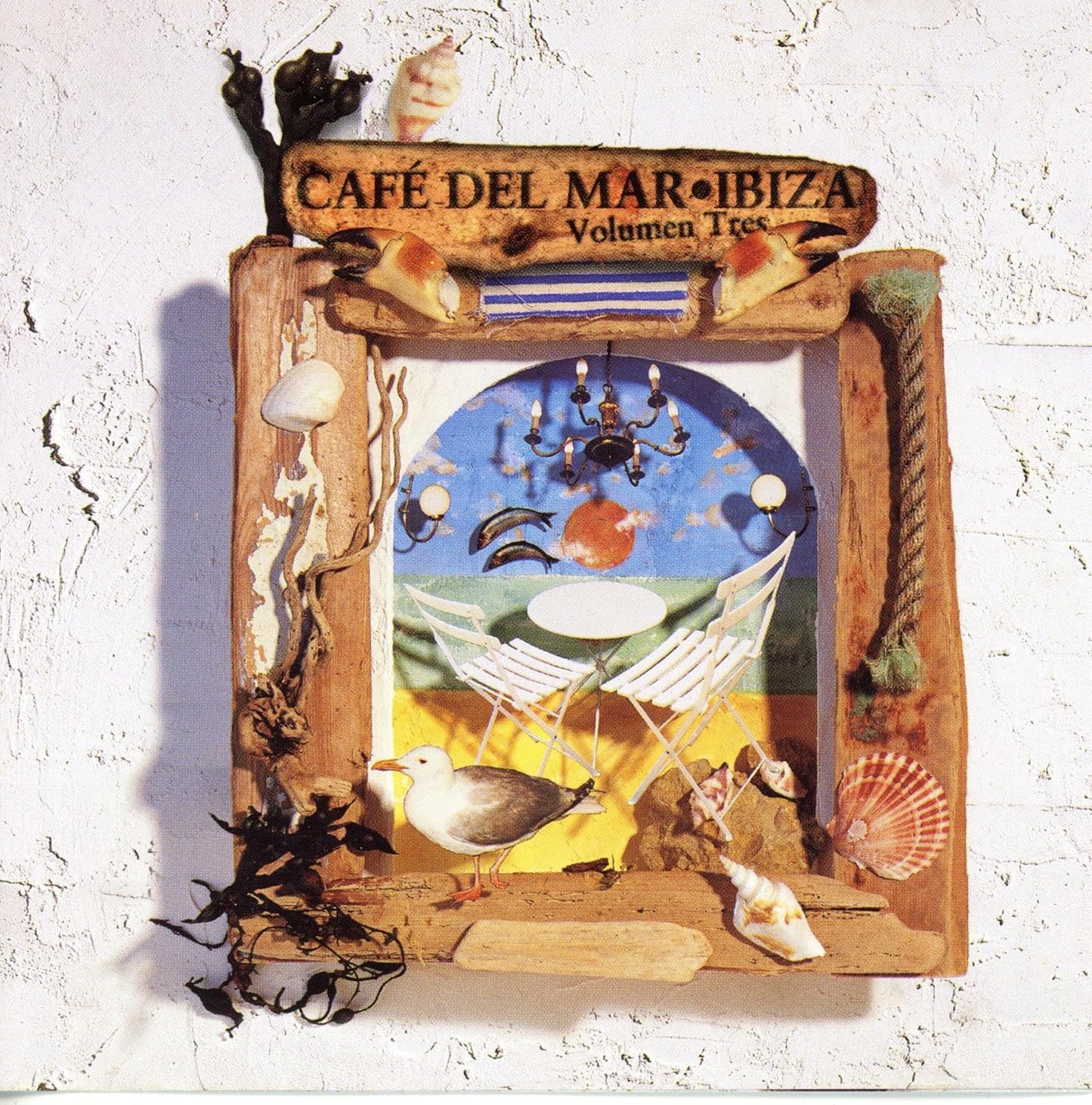 A framed decorative artwork mounted on a white textured wall. The piece features a rustic wooden frame adorned with shells, rope, seaweed, and small seagull figures, surrounding a colorful central illustration with hammocks, a chandelier, and the text 'CAFÉ DEL MAR IBIZA Volumen Tree'.