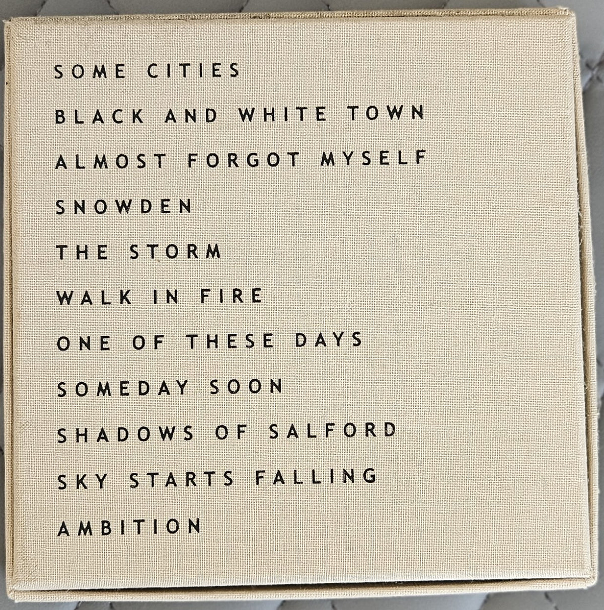 A square canvas wall art print with a beige linen-textured background and black printed text listing song titles, photographed against a light marble surface.