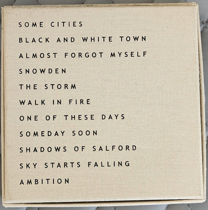 A square canvas wall art print with a beige linen-textured background and black printed text listing song titles, photographed against a light marble surface.