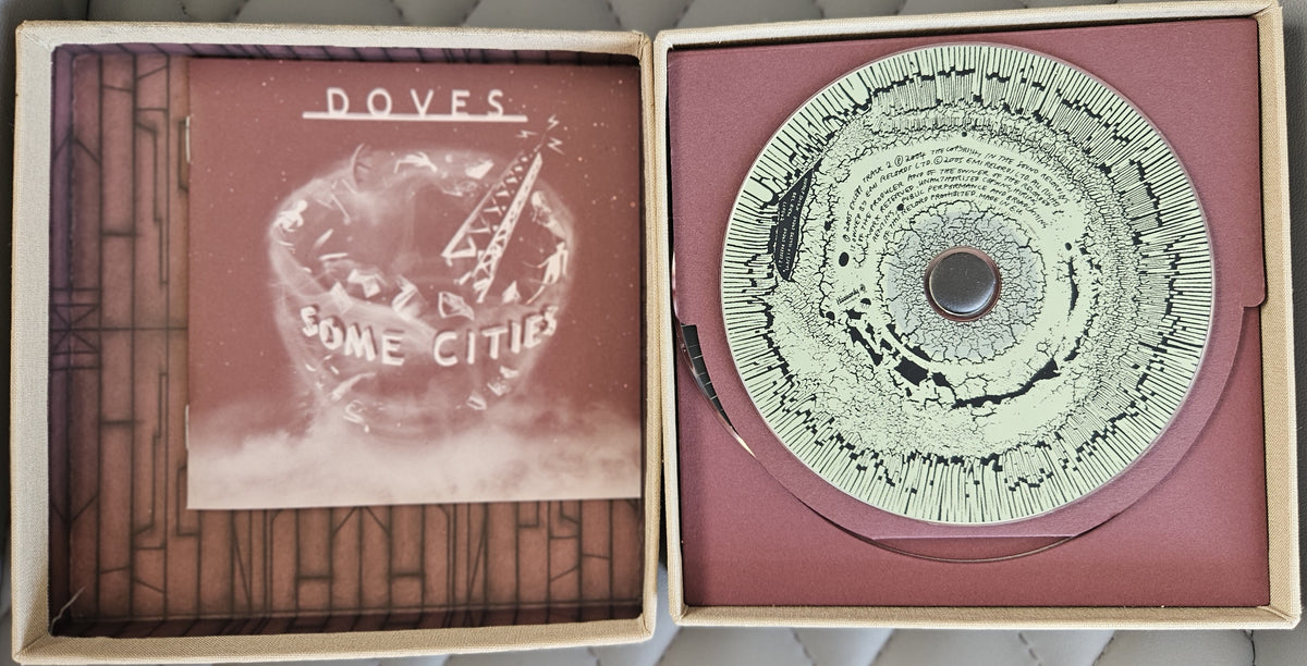 An open cardboard record sleeve display showing a vinyl record and printed insert. The left panel contains a printed insert with the text 'DOVES' and 'SOME CITY', and the right panel holds a circular record with a dark center and fine printed pattern on a dusty rose background.