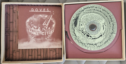 An open cardboard record sleeve display showing a vinyl record and printed insert. The left panel contains a printed insert with the text 'DOVES' and 'SOME CITY', and the right panel holds a circular record with a dark center and fine printed pattern on a dusty rose background.