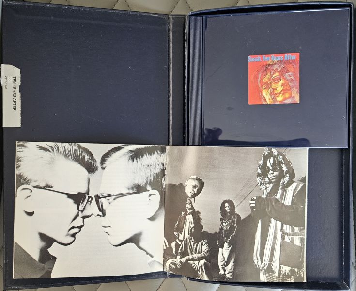 A vintage music album package opened flat, showing a dark blue sleeve with a small red cover insert, black-and-white band photos printed on the front, and a white paper tag attached to the left side.