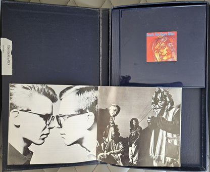 A vintage music album package opened flat, showing a dark blue sleeve with a small red cover insert, black-and-white band photos printed on the front, and a white paper tag attached to the left side.