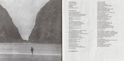 An open book spread showing a black-and-white photograph of a person walking between large rocky cliffs on the left page and printed song lyrics on the right page.